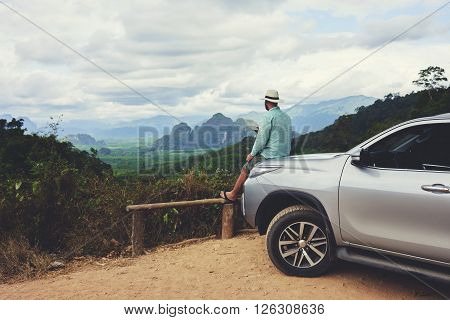 Young man wanderer is enjoying beautiful landscape during summer adventure on rental automobile in Asia. Male traveler is dreaming about something while is sitting on a car hood against Amazon view