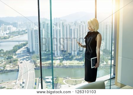 Confident fyoung emale financier with touch pad in hand is reading financial news in network via mobile phonewhile is standing near private office window with view of high buildings business district