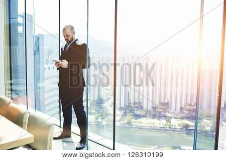 Businessman is searching information on web page via mobile phone while is standing in his private office near window with view of metropolitan city with tall skyscrapers background with copy space