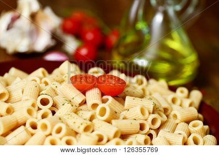 closeup of a plate with cooked penne rigate on a table with garlic cloves, tomatoes and a glass cruet with olive oil in the background