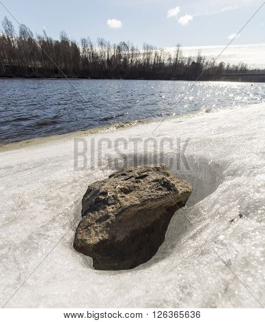 Stone in Ice by River with trees and a patrly cloudy sky