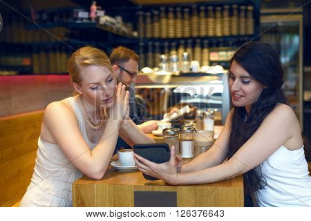 Two young women looking at a digital tablet as they sit enjoying coffee together in a bistro or coffeebar