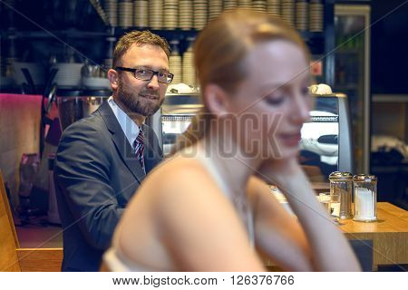 Middle-aged businessman with eyeglasses looking at the camera while sitting in a crowded coffeebar