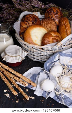 Basket with freshly baked pastries, bread sticks, milk, coffee and jam on wooden table.