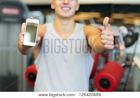 sport, bodybuilding, lifestyle, technology and people concept - close up of happy young man showing thumbs up and smartphone blank screen in gym