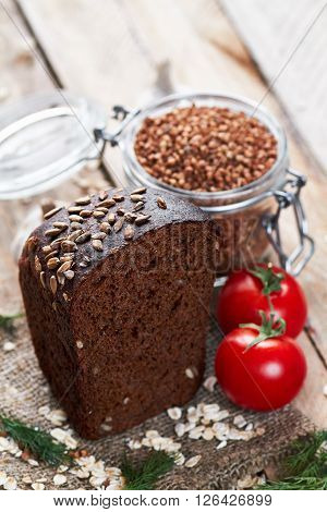 Close up photo of rustic bread sprinkled with sunflower seeds on sackcloth, two tomatoes, dill, oat flakes and jar with buckwheat on planked wooden table