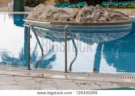 Metal handhold entrance to the blue hotel pool with stone island in a middle of the pool with reflection in water