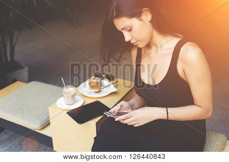 Attractive hipster girl is chatting with her friend on moile phone while is waiting for the order in cafe. Woman is searching information in network by using cell telephone during lunch in work break