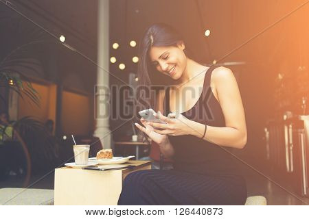 Attractive female student is reading laughable article in network on mobile phone while is sitting in university cafe. Woman chatting with her friends by cell telephone during breakfast in coffe shop