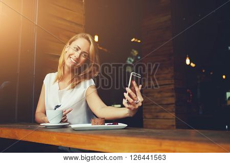Charming happy hipster girl photographing herself on cell telephone camera while relaxing in modern coffee shop pretty woman with cute smile making self photo on her smart phone during rest in cafe