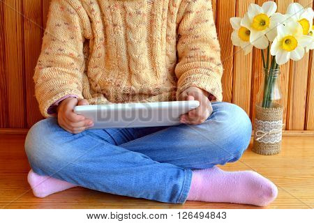Child sits holding a tablet in hands. Vase of daffodils. Wooden background