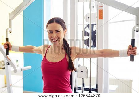 Pretty Caucasian smiling woman is working out on butterfly machine in gym