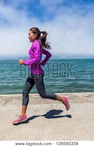 Woman running on waterfront harbour pier in San Francisco, California, USA. Mixed race fit fitness sport model in spring / autumn running clothing outfit. Smiling happy female athlete runner training.