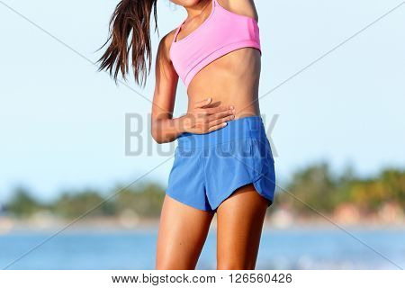 Side stitch stomach pain. Woman runner cramps. Jogging girl with stomach side pain during jogging work out. Female athlete on beach.