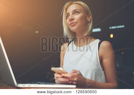 Young beautiful blonde female holding mobile phone while sitting with portable net-book in coffee shop interior charming dreamy woman using cell telephone and laptop computer during rest in cafe