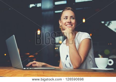 Young female with cute smile sitting with portable net-book in modern coffee shop interior during recreation time charming happy woman student using laptop computer to prepare for the course work