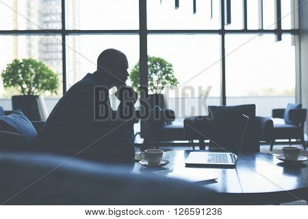 Silhouette of a businessman is thinking about future conversations with investors while is sitting in cafe with net-book. Male financier is watching financial news in internet via laptop computer