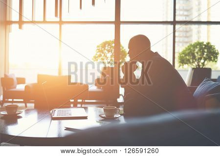 Silhouette of man managing director is thinking how to prevent the company's crisiswhile is sitting with net-book in cafe. Male financier in watching world news on laptop computer during coffee break