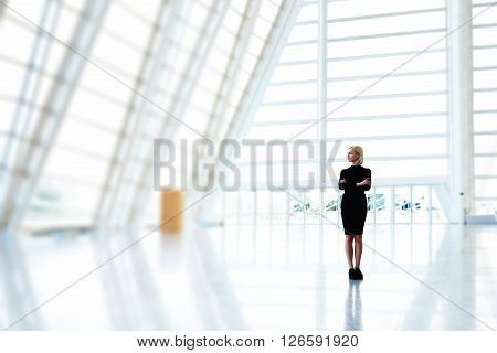 Young female property buyer is thinking of buying a modern skyscraper premises for future office space. Woman entrepreneur is standing with crossed arms in empty interior with copy space for advertise