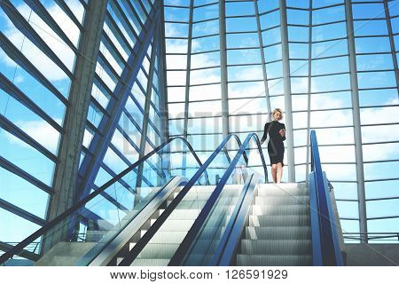 Young businesswoman is searching information on website via cell telephonewhile is standing near escalator staircase in modern airport interior with contemporary building design Copy space background
