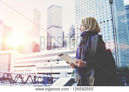 Young beautiful woman with portable touch pad in hands is enjoying view of Hong Kong business center outside the window background with copy space for advertising text message or promotional content