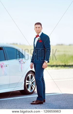 Portait of a young handsome man in suite near white widding decorated car.  Gorgeous guy, outdoors