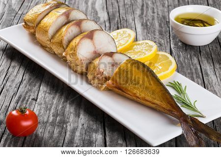 Slices of Smoked fish Mackerel or Scomber on a white rectangular dish with cherry tomato sliced lemon and rosemary on an old wooden table small bowl with olive oil studio lights top view