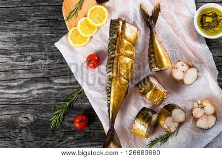 Slices of Smoked fish Mackerel or Scomber on a white parchment paper with cherry tomato sliced lemon and rosemary on an old wooden table with small bowl with olive oil studio ligths top view