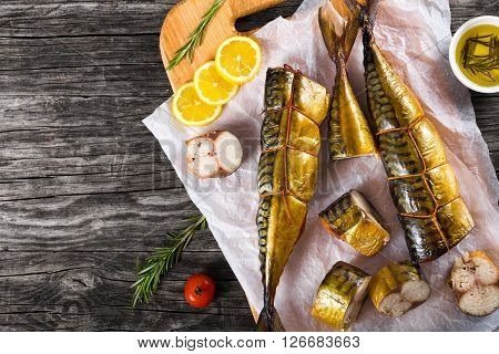 Slices of Smoked fish Mackerel or Scomber on a white parchment paper with cherry tomato sliced lemon and rosemary on an old wooden table with small bowl with olive oil studio ligths top view