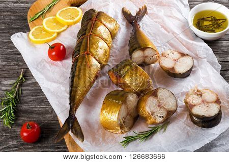 Slices of Smoked fish Mackerel or Scomber on a white parchment paper with cherry tomato sliced lemon and rosemary on an old wooden table with small bowl with olive oil studio ligths top view