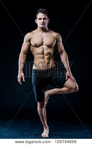 Muscular man balancing on the one leg feeling relaxed on dark background