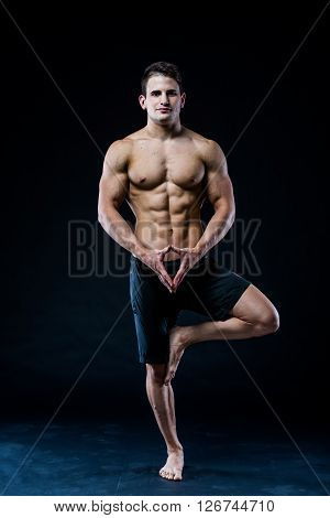 Muscular man balancing on the one leg feeling relaxed on dark background