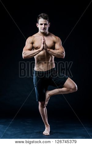 Muscular man balancing on the one leg on dark background