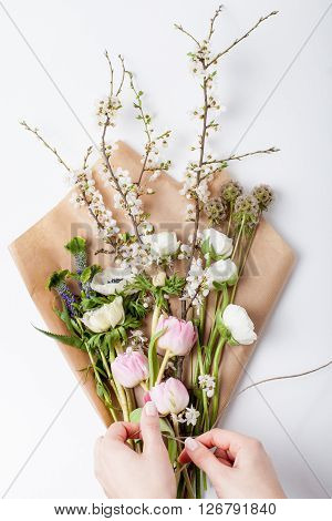 Hands packing flowers: pink tulips white anemones white buttercups and blossom plum tree branch lying on wrapping paper from the top