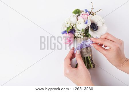Two hands of the florist are making a bow from a ribbon in beautifull spring bouquet from pink tulips violet grape hyacinths white anemones violet veronica and white buttercups on white background