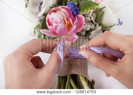 Two woman hands are making a bow from a ribbon in beautiful spring bouquet from pink tulips violet grape hyacinths white anemones violet veronica and white buttercups on white background