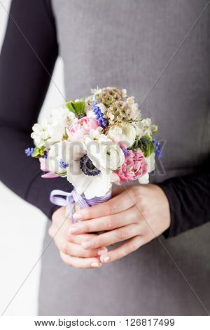 Woman in grey dress and black shirt is holding a spring bouquet from pink tulips violet grape hyacinths white anemones violet veronica and white buttercup with violet ribbon