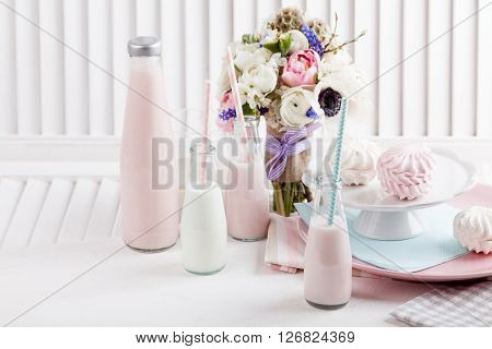 Four bottles of milkshakes three with straws and glass of milk and plates with pink and white meringues and cloth on white wooden shutter with bouquet from pink tulips violet grape hyacinths white anemones violet veronica and white buttercup with violet r