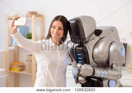 Say cheese. Cheerful beautiful  nice woman holding cellphone and making selfies  with the robot