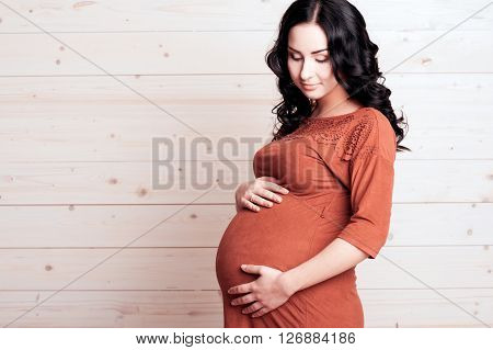 Smiling pregnant woman 20-24 year old holding tummy over wooden background. Wearing stylish dress closeup. Maternity.