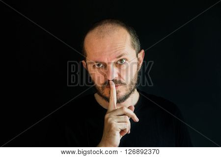 Serious man with beard and mustaches on black background in low key, making silence gesture, pst, shh