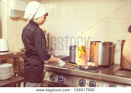 Chef Cooking With Fire In Frying Pan. Professional chef in a commercial kitchen cooking flambe style. Chef frying food in flaming pan on gas hob in commercial kitchen.