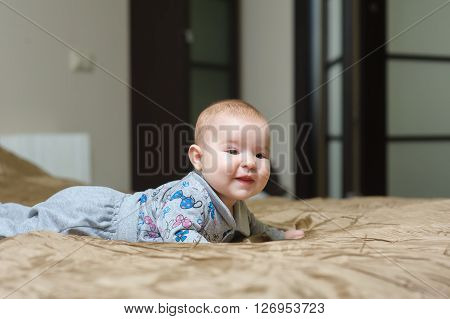Baby girl lying on belly on bed in bedroom with golden linens. Charming 5-month-old girl smiling and grimacing. Home interior in background.
