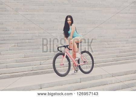 Sexy girl on bicycle. Young slim sexy sportive woman in blue shorts and white snickers long-haired, sensual posing on pink fix bicycle at grey stairs background. Soft toned.