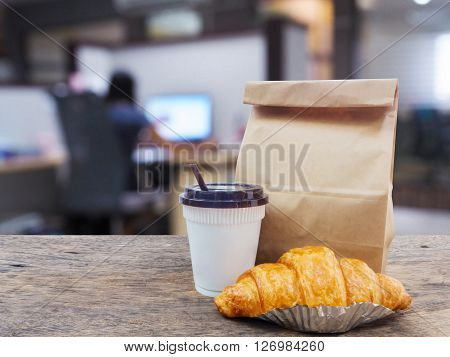 coffee and croissant with paper bag on wooden table over office blurred background