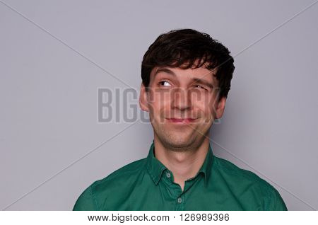 leer European man in a green shirt on a neutral gray background. Dodgy emotion. Look up to the left with one eye half-closed