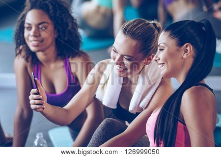 I can see progress in my workouts! Beautiful young women in sportswear discussing something with smile and using smartphone while sitting on exercise mat at gym