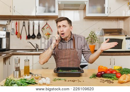 Perturbed Stress Young Man In Apron Sitting At Table With Vegetables, Talking On Mobile Phone, Cooki