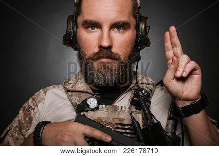 Close-up Portrait Of Brutal Bearded Man In Military Desert Uniform And Body Armor Who Holds His Gun