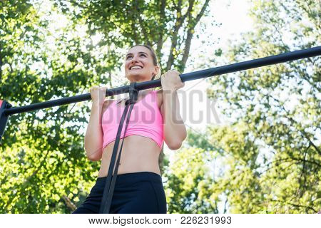 Low-angle view of a fit and cheerful young woman, wearing pink sports bra while doing chin-up exercise for the upper-body outdoors in a calisthenics park